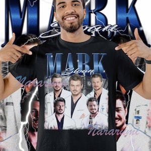 Puede incluir: Camiseta negra con texto "MARK Sloan" en azul estilizado e im&aacute;genes de actores con batas blancas. El dise&ntilde;o incluye un fondo azul con rayos. La camiseta es de cuello redondo.