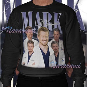 May include: Black sweatshirt featuring a collage of images and the name "MARK Sloan" in a purple retro font. The sweatshirt has a crew neck and long sleeves. The word "Nararumi" is printed on the bottom right.