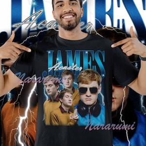Puede incluir: Camiseta negra con un gráfico de James Acaster, con el texto "JAMES Acaster" en azul. El diseño incluye múltiples imágenes de James Acaster y la palabra "Nararumi" en rosa. El fondo tiene un diseño de rayo azul.