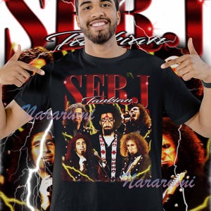 May include: Black t-shirt featuring a graphic of Serj Tankian and other band members. The design includes the band's name in red, with a background of lightning bolts and a blurred image of the band members. The shirt is worn by a person.