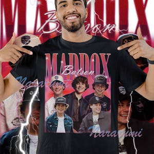 Puede incluir: Camiseta negra con un diseño retro que presenta el nombre "MADDOX Batson" y un grupo de jóvenes. El diseño tiene un aspecto vintage descolorido con tonos rosas y rojos. El fondo es una imagen borrosa.