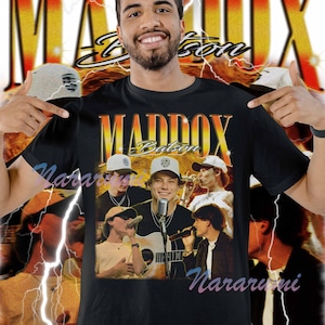 Puede incluir: Camiseta negra con el texto "MADDOX Batsen" en naranja y amarillo de estilo vintage. La camiseta tiene un gráfico de una banda. El fondo tiene un diseño de relámpagos.