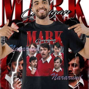 May include: Black t-shirt featuring a graphic of Mark Corrigan in a red sweater and white collar. The text "MARK Corrigan" is displayed in red, with a lightning bolt design in the background. The shirt is worn by a person.