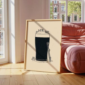 Puede incluir: Una impresión artística enmarcada presenta una ilustración caprichosa de una gran pinta de cerveza oscura con espuma. Se representan pequeñas figuras subiendo escaleras y de pie sobre la espuma. La obra de arte es en blanco y negro sobre un fondo beige.