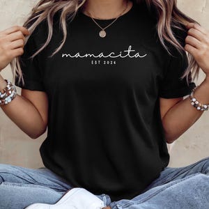 Könnte beinhalten: Schwarzes T-Shirt mit dem kursiven Text "mamacita" und dem Text "EST 2026" in Weiß. Das Shirt hat einen Rundhalsausschnitt und wird mit blauen Jeans und weißen Turnschuhen getragen. Eine goldene Halskette und Armbänder sind ebenfalls sichtbar.