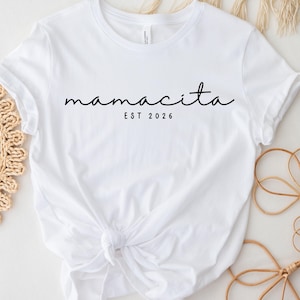 Könnte beinhalten: Ein weißes T-Shirt mit dem Wort "mamacita" in Schreibschrift und "EST 2026" darunter. Das T-Shirt ist an der Taille geknotet. Dekorative Elemente sind im Hintergrund zu sehen.