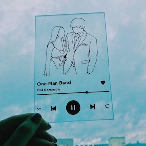 May include: A glass print with a black and white illustration of a couple. The print has a Spotify-like design with the song title "One Man Band" by Old Dominion.