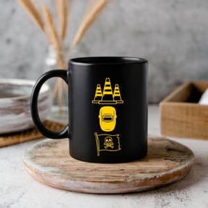 May include: Black ceramic mug with a yellow graphic of three traffic cones, a welding mask, and a pirate flag with a skull and crossbones. The mug is on a wooden coaster.