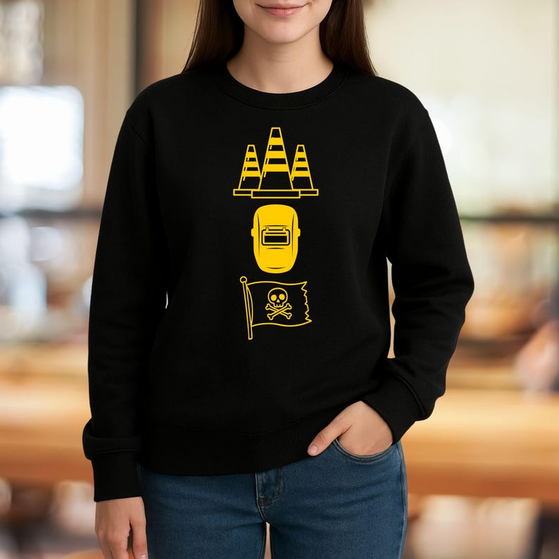 May include: Black sweatshirt with yellow graphic of three traffic cones, a welding mask, and a pirate flag with a skull and crossbones. The sweatshirt is made of a soft material.