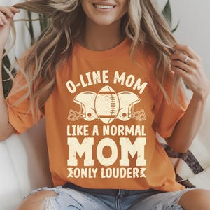 Puede incluir: Camiseta naranja con un gráfico blanco que dice "O-Line Mom Like A Normal Mom Only Louder" con un balón de fútbol y dos cascos.