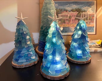 Puede incluir: Cuatro árboles decorativos en forma de cono construidos con piezas de vidrio marino en tonos azules y turquesas. Cada árbol está montado sobre una base circular de rodaja de madera y rematado con una estrella de mar blanca.