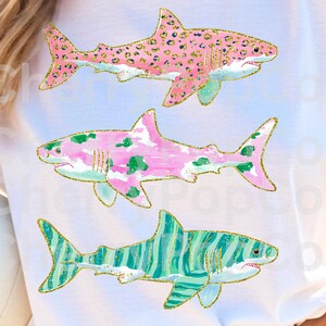 May include: White t-shirt featuring three colorful shark illustrations. The sharks have pink, green, and blue patterns with gold glitter outlines. The top shark has a pink leopard print, the middle shark has pink and green spots, and the bottom shark has blue and green stripes.