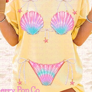May include: A pale yellow t-shirt featuring a graphic print of a bikini made from iridescent pink and teal seashells. The bikini top and bottom are accented with strings of white pearls, small pink starfish, and delicate bows at the shoulders.