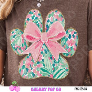 Puede incluir: Una camiseta marr&oacute;n con un dise&ntilde;o de huella de pata y un lazo rosa. La huella de la pata est&aacute; llena de un estampado floral en rosa, verde y azul, delineado con purpurina dorada. El texto "CHERRY POP CO" y "PNG DESIGN" est&aacute; en la parte inferior.