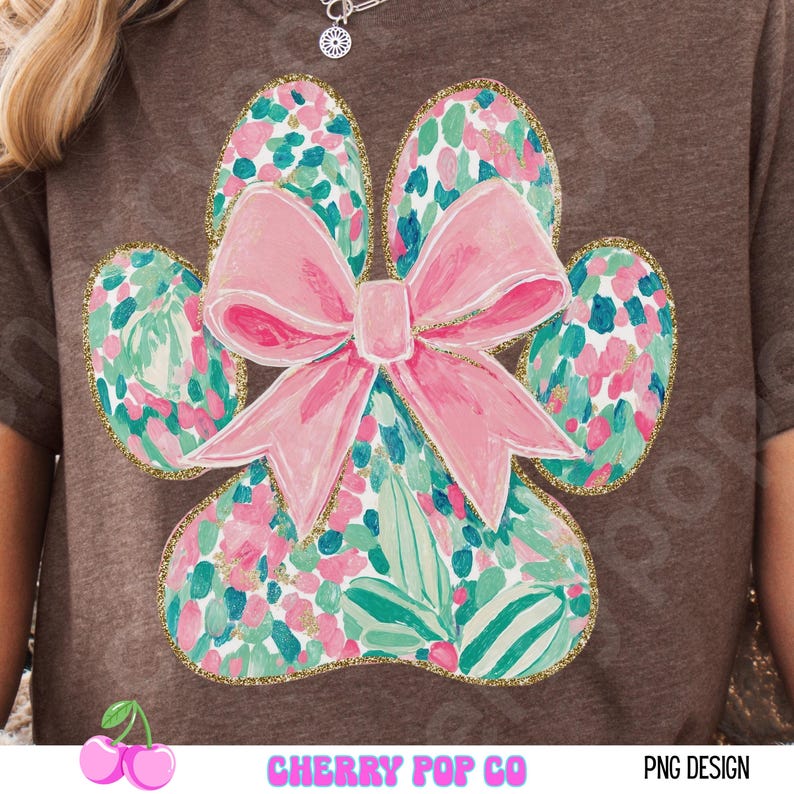 Puede incluir: Una camiseta marr&oacute;n con un dise&ntilde;o de huella de pata y un lazo rosa. La huella de la pata est&aacute; llena de un estampado floral en rosa, verde y azul, delineado con purpurina dorada. El texto "CHERRY POP CO" y "PNG DESIGN" est&aacute; en la parte inferior.