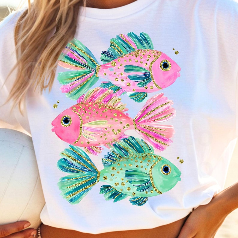 May include: White t-shirt featuring a colorful illustration of three fish. The fish have pink, teal, and gold accents. The fish are arranged vertically, with a playful and whimsical design.