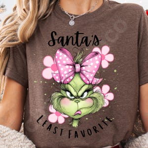 Puede incluir: Camiseta marr&oacute;n con un dise&ntilde;o del Grinch de dibujos animados. El Grinch es verde con una expresi&oacute;n enfadada, un lazo rosa con lunares y flores rosas. El texto dice "Santa's Least Favorite".
