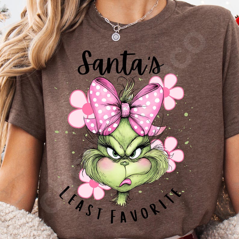 Puede incluir: Camiseta marr&oacute;n con un dise&ntilde;o del Grinch de dibujos animados. El Grinch es verde con una expresi&oacute;n enfadada, un lazo rosa con lunares y flores rosas. El texto dice "Santa's Least Favorite".