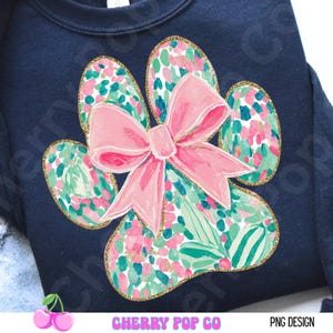 Puede incluir: Sudadera azul marino con un dise&ntilde;o de gran huella de pata y un lazo rosa. La huella de la pata est&aacute; llena de un estampado floral en rosa, verde y blanco, delineado con purpurina dorada. El texto "CHERRY POP CO" y "PNG DESIGN" est&aacute; en la parte inferior.