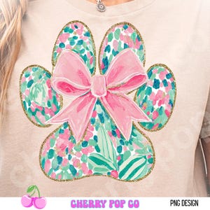 Puede incluir: Camiseta beige claro con un dise&ntilde;o de huella de pata. La huella est&aacute; decorada con patrones florales rosas, turquesas y verdes y delineada con purpurina dorada. Un gran lazo rosa est&aacute; centrado en la huella. El texto "CHERRY POP CO" est&aacute; en la parte inferior.