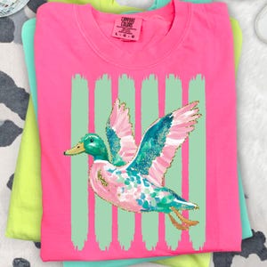 K&ouml;nnte beinhalten: Ein pinkfarbenes Comfort Colors T-Shirt mit einem bunten Enten-Design mit t&uuml;rkisfarbenen, rosa und goldenen Akzenten. Die Ente ist vor einem Hintergrund aus vertikalen mintgr&uuml;nen Streifen platziert. Das T-Shirt ist gefaltet und mit anderen Shirts gestapelt.