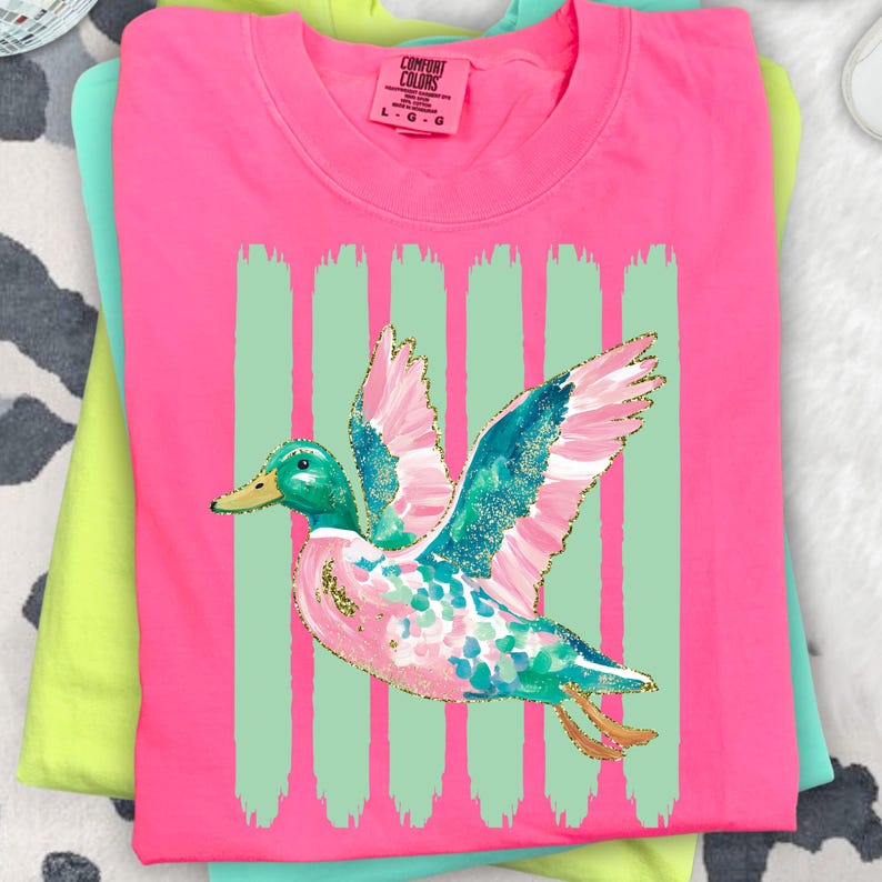 K&ouml;nnte beinhalten: Ein pinkfarbenes Comfort Colors T-Shirt mit einem bunten Enten-Design mit t&uuml;rkisfarbenen, rosa und goldenen Akzenten. Die Ente ist vor einem Hintergrund aus vertikalen mintgr&uuml;nen Streifen platziert. Das T-Shirt ist gefaltet und mit anderen Shirts gestapelt.