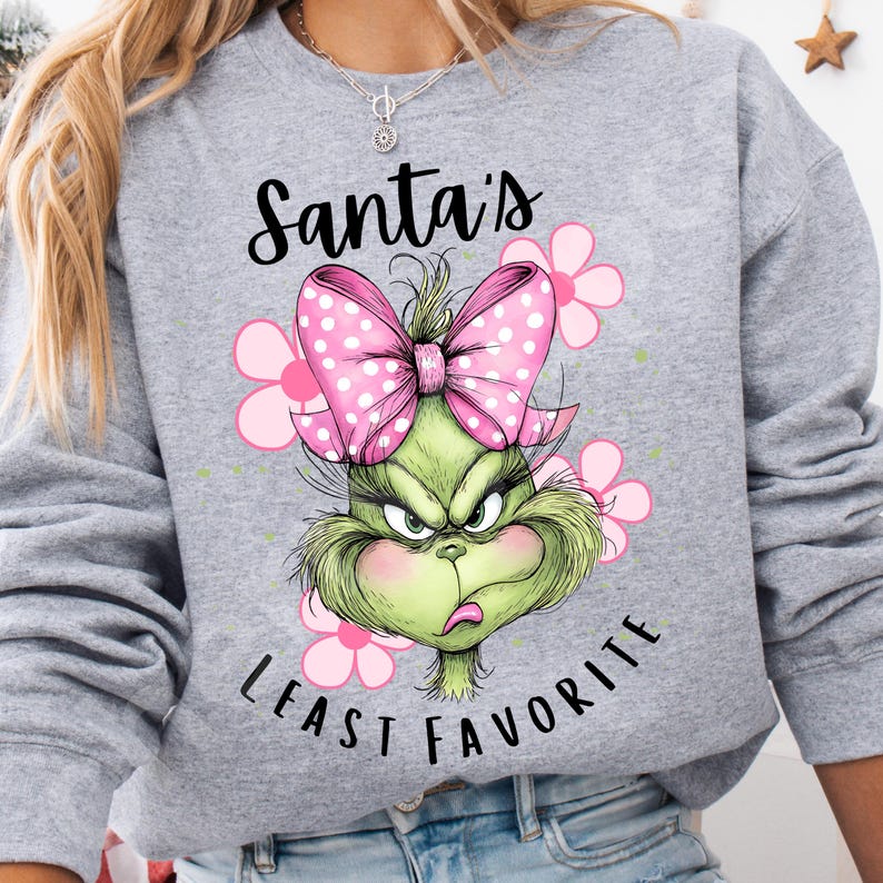 Puede incluir: Una sudadera gris con un dise&ntilde;o del Grinch, un lazo rosa y flores rosas. El texto "Santa's Least Favorite" est&aacute; impreso debajo del Grinch. El dise&ntilde;o es un gr&aacute;fico festivo con tem&aacute;tica navide&ntilde;a.