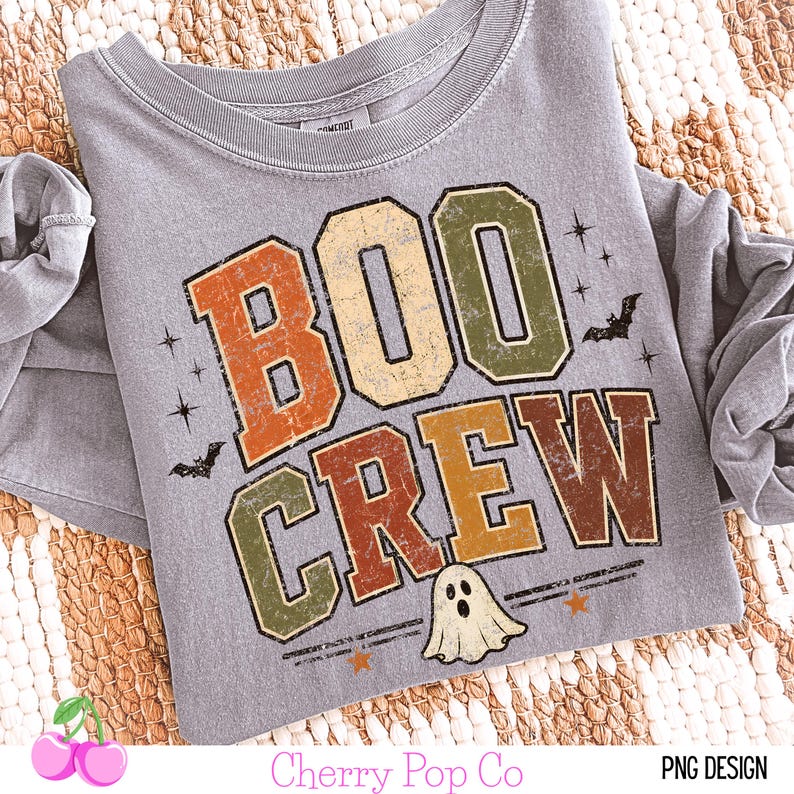 May include: A grey long-sleeve t-shirt featuring the text BOO CREW in distressed orange, cream, and olive green block letters. The design includes small black bats, stars, and a white ghost graphic with a surprised expression at the bottom.