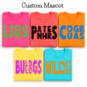 Op de afbeelding: Vijf gevouwen T-shirts met ronde hals in felle kleuren. Het roze shirt heeft groene tekst met LIONS. Het koraalkleurige shirt heeft zwarte tekst met PANTHERS. Het oranje shirt heeft blauwe tekst met COUGARS. Het gele shirt heeft paarse tekst met BULLDOGS. Het turquoise shirt heeft oranje tekst met WILDCATS.