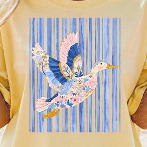 May include: A light yellow t-shirt featuring a colorful duck design. The duck has pink, blue, and yellow accents, with floral patterns on its body and wings. The background has vertical blue stripes.