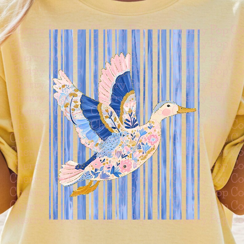 May include: A light yellow t-shirt featuring a colorful duck design. The duck has pink, blue, and yellow accents, with floral patterns on its body and wings. The background has vertical blue stripes.
