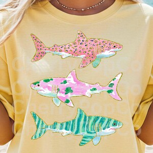 K&ouml;nnte beinhalten: Gelbes T-Shirt mit drei bunten Hai-Illustrationen. Die Haie haben rosa, gr&uuml;ne und blaue Akzente mit goldenen Glitzerdetails. Der obere Hai hat ein rosa Leopardenmuster.
