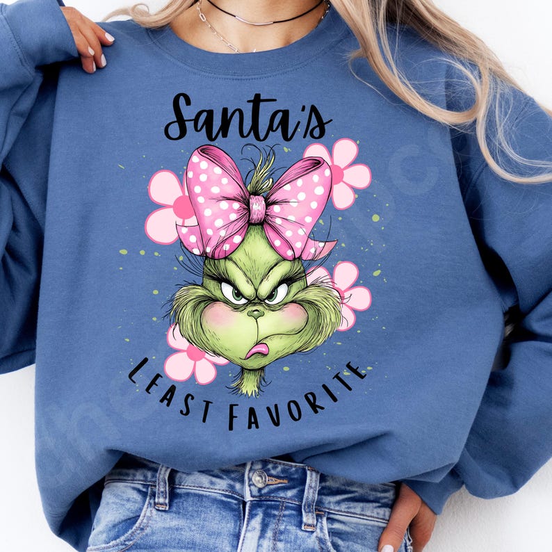 Puede incluir: Sudadera azul con un dise&ntilde;o del Grinch, un lazo rosa y flores. El texto dice "Santa's Least Favorite". El Grinch es verde con una expresi&oacute;n enfadada. Un dise&ntilde;o gr&aacute;fico divertido y festivo.