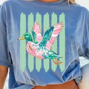 K&ouml;nnte beinhalten: Hellblaues T-Shirt mit einem farbenfrohen Enten-Design. Die Ente hat rosa, t&uuml;rkis und goldfarbene Akzente, einen gr&uuml;nen Kopf und einen gelben Schnabel. Vertikale gr&uuml;ne Streifen befinden sich hinter der Ente.