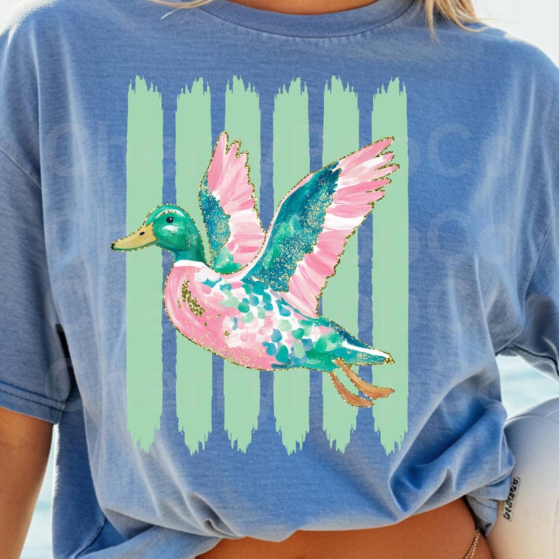 K&ouml;nnte beinhalten: Hellblaues T-Shirt mit einem farbenfrohen Enten-Design. Die Ente hat rosa, t&uuml;rkis und goldfarbene Akzente, einen gr&uuml;nen Kopf und einen gelben Schnabel. Vertikale gr&uuml;ne Streifen befinden sich hinter der Ente.