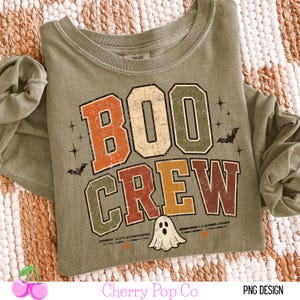 May include: An olive green long-sleeve shirt featuring the text BOO CREW in a distressed, multi-colored block font. The design includes small black bat silhouettes, starburst accents, and a white ghost illustration centered below the text.