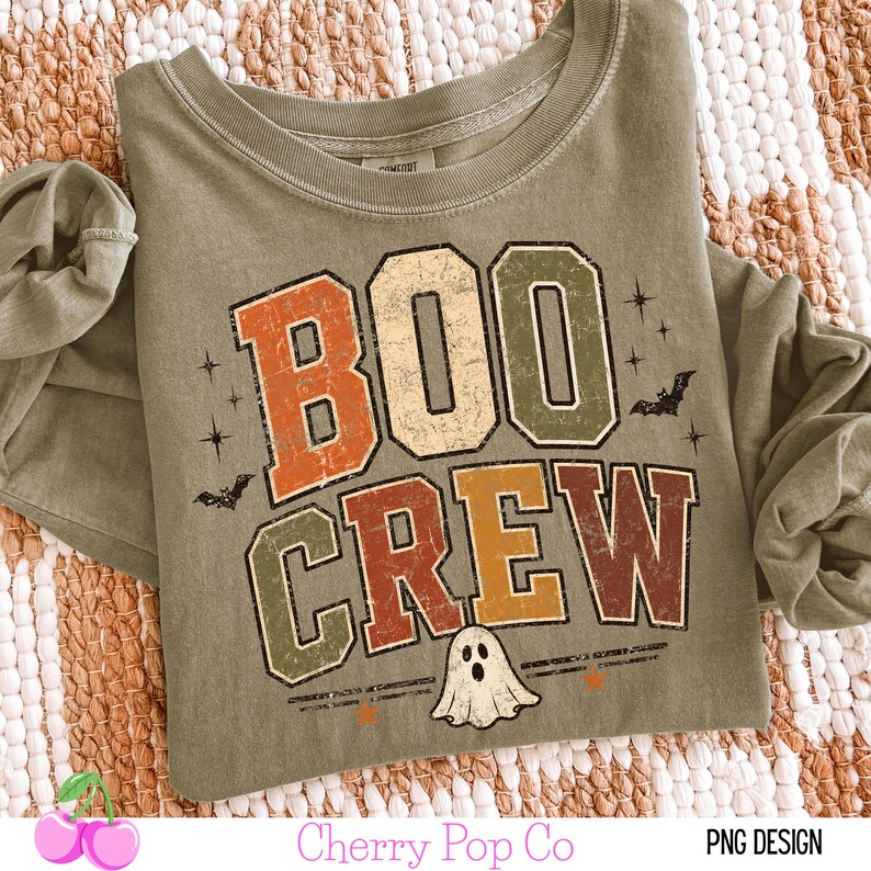 May include: An olive green long-sleeve shirt featuring the text BOO CREW in a distressed, multi-colored block font. The design includes small black bat silhouettes, starburst accents, and a white ghost illustration centered below the text.