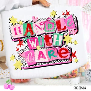 Puede incluir: Bolso tote blanco con las palabras "HANDLE WITH CARE" en letras de bloque coloridas. El diseño incluye detalles en rosa, verde y rojo, con adornos de estrellas y corazones. El bolso es sostenido por una persona.