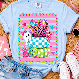 May include: A light blue t-shirt featuring a graphic of three stacked pumpkins with a large colorful bow on top. The pumpkins have leopard, checkered, and polka dot patterns. A small white ghost and the word BOO are printed in the design.