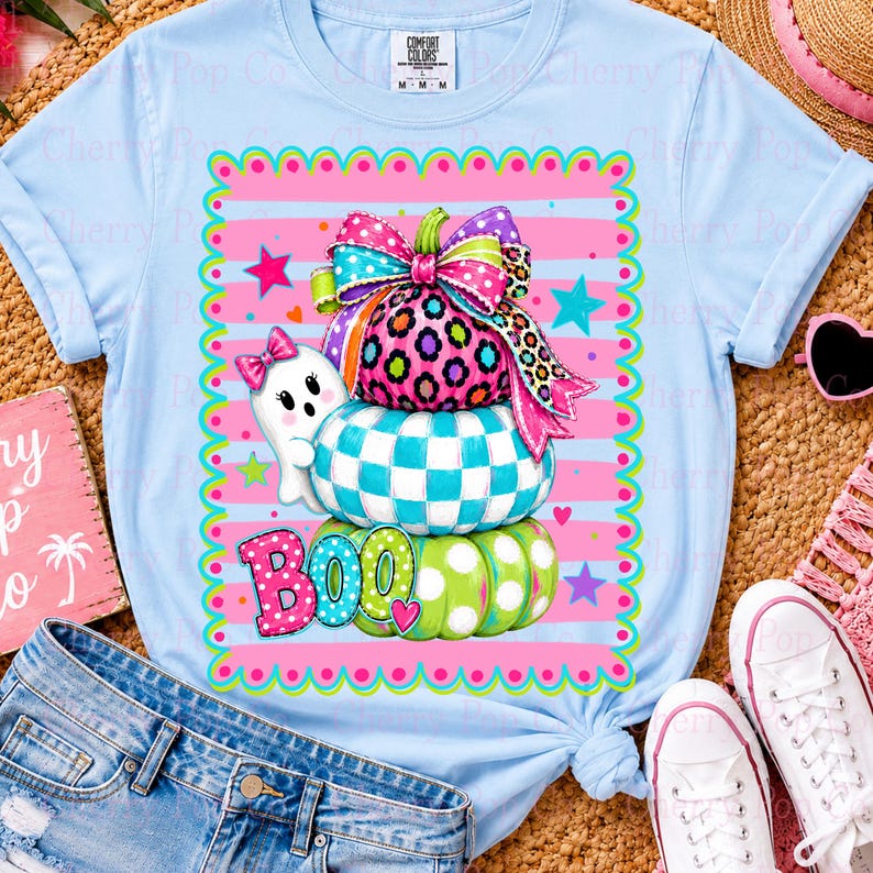 May include: A light blue t-shirt featuring a graphic of three stacked pumpkins with a large colorful bow on top. The pumpkins have leopard, checkered, and polka dot patterns. A small white ghost and the word BOO are printed in the design.