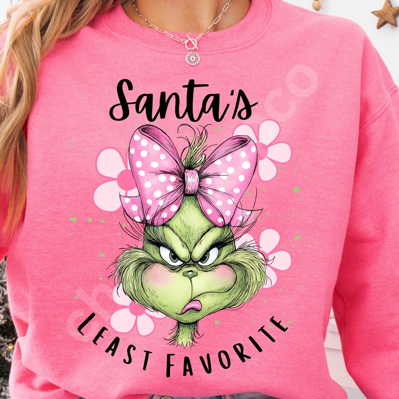 Puede incluir: Sudadera rosa con un gr&aacute;fico del Grinch, un lazo rosa con lunares y el texto "Santa's Least Favorite". El Grinch es verde con una expresi&oacute;n gru&ntilde;ona. Flores blancas y rosas en el fondo.