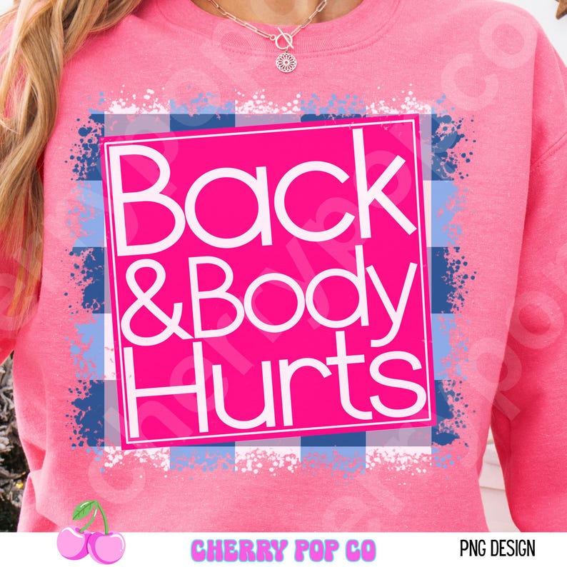 May include: Pink sweatshirt with the text "Back & Body Hurts" in a white font, set against a pink background. The text is inside a white square with a blue and white plaid pattern. The sweatshirt is a crew neck.