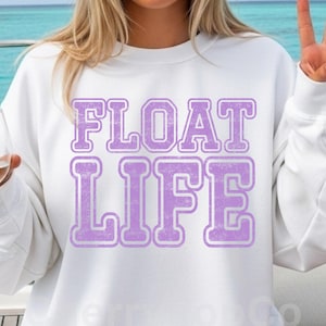May include: White sweatshirt with the words "FLOAT LIFE" in large, lavender block letters. The text has a distressed, vintage look. The sweatshirt is a casual, comfortable style.