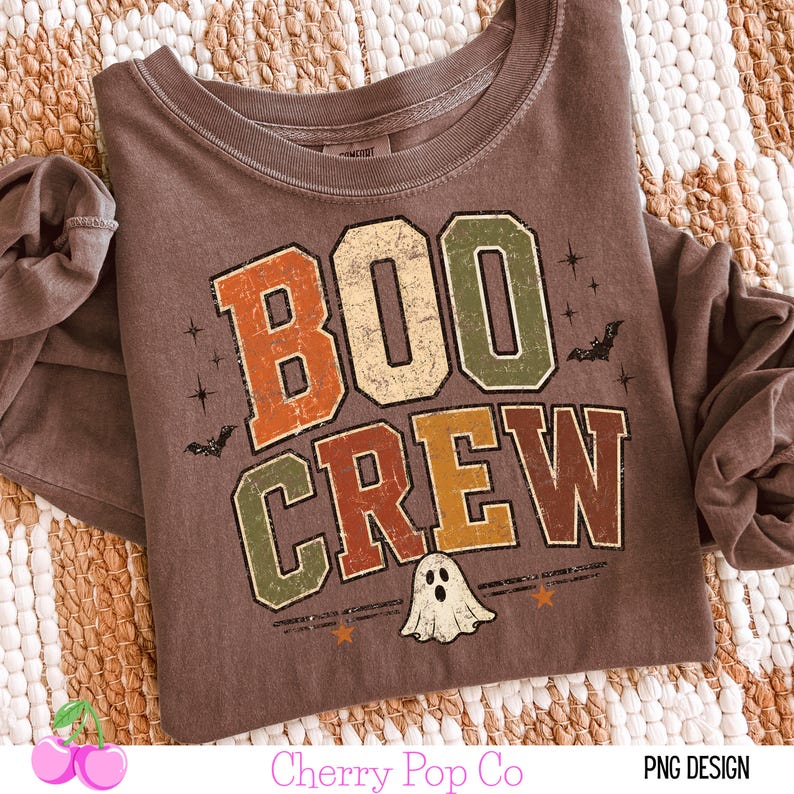 May include: A brown long-sleeve t-shirt featuring the text BOO CREW in distressed block letters colored in orange, green, and brown. The graphic includes small black bats, star shapes, and a white ghost illustration centered below the text.