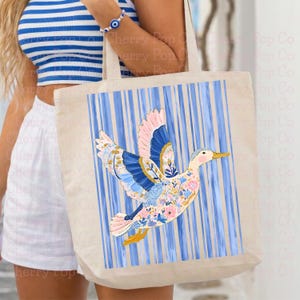 May include: Beige tote bag with a blue and white striped background and a colorful duck illustration. The duck features pink, blue, and floral patterns. The bag has a rectangular shape and two handles.