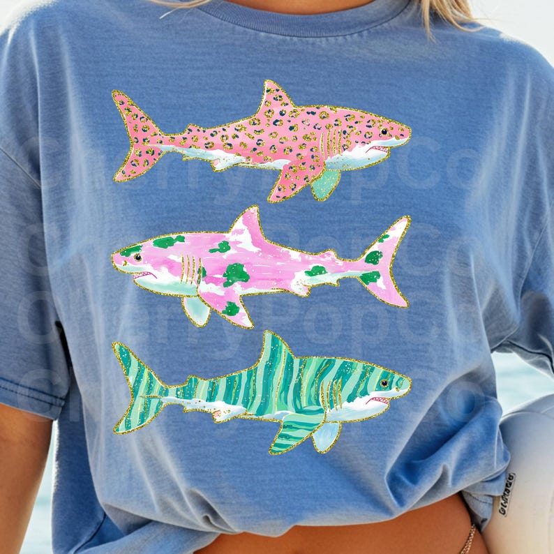 K&ouml;nnte beinhalten: Hellblaues T-Shirt mit drei stilisierten Hai-Illustrationen. Die Haie haben rosa, gr&uuml;ne und blaue Muster mit goldenen Glitzerakzenten. Der obere Hai hat einen rosa Leopardenmuster, der mittlere Hai hat gr&uuml;ne Flecken und der untere Hai hat blaue Streifen.