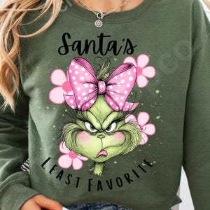 Puede incluir: Sudadera verde oliva con un dise&ntilde;o del Grinch, un lazo rosa con lunares y flores rosas. El texto dice "Santa's Least Favorite". El dise&ntilde;o est&aacute; centrado en la parte delantera.