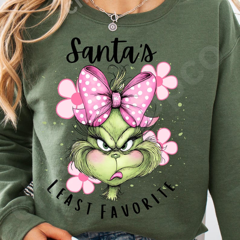 Puede incluir: Sudadera verde oliva con un dise&ntilde;o del Grinch, un lazo rosa con lunares y flores rosas. El texto dice "Santa's Least Favorite". El dise&ntilde;o est&aacute; centrado en la parte delantera.