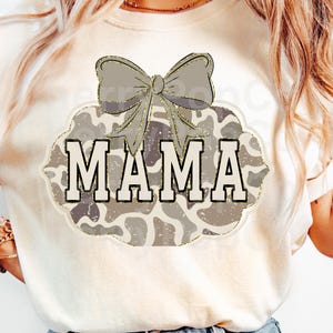 以下が含まれることがあります： クリーム色のTシャツ。デザインは、太字の白い文字で「MAMA」という言葉、灰色のリボン、金色のグリッターで縁取られた迷彩柄が特徴です。