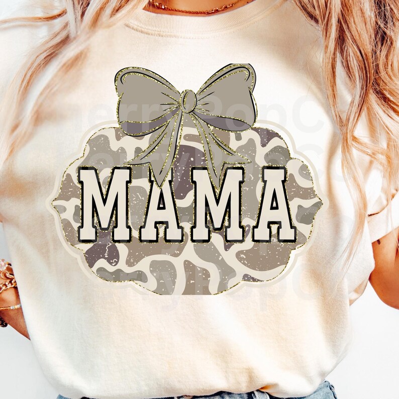 以下が含まれることがあります： クリーム色のTシャツ。デザインは、太字の白い文字で「MAMA」という言葉、灰色のリボン、金色のグリッターで縁取られた迷彩柄が特徴です。